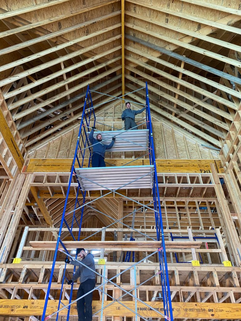 Three smiling electricians stand on a very tall scaffolding structure in a  housing frame